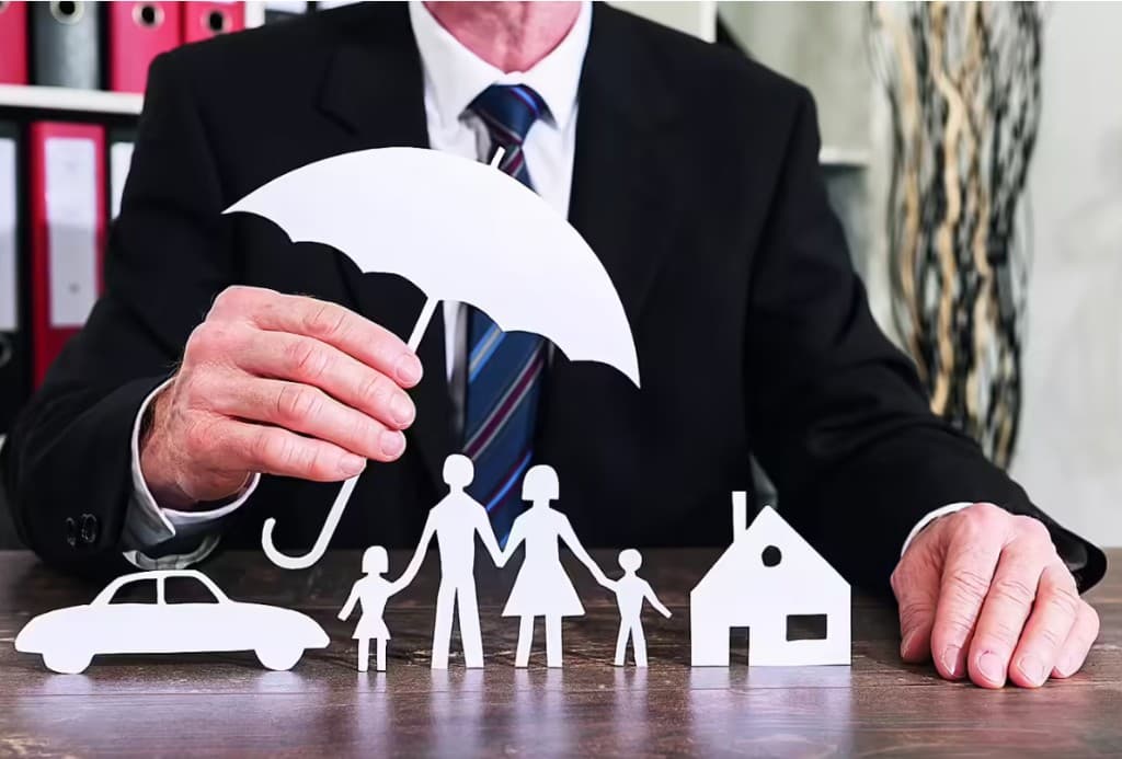 Insurance concept: a professional holding a paper umbrella over cutouts representing a car, a family, and a home.